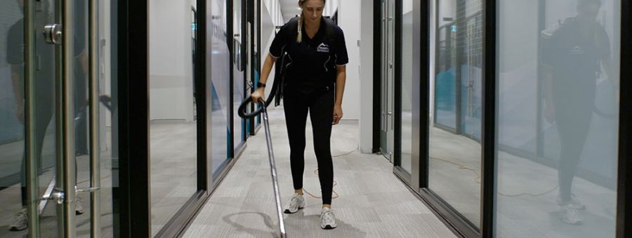 Urban Clean staff member vacuuming a carpeted corridor in an office building.